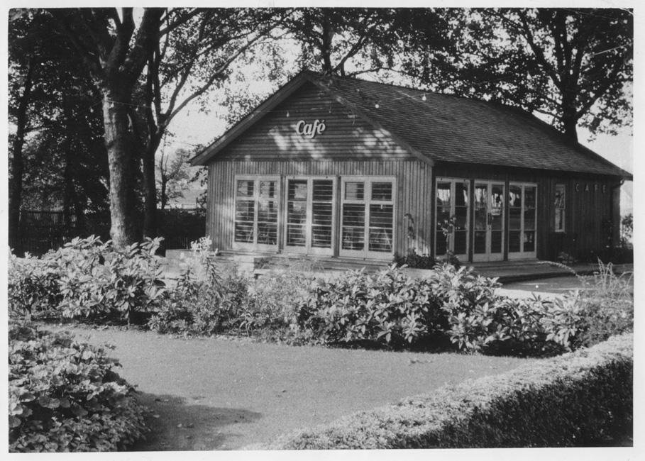 Black and white image of a simple building labelled, ‘Cafe,’ surrounded by trees. In front are well kept beds and small hedges.