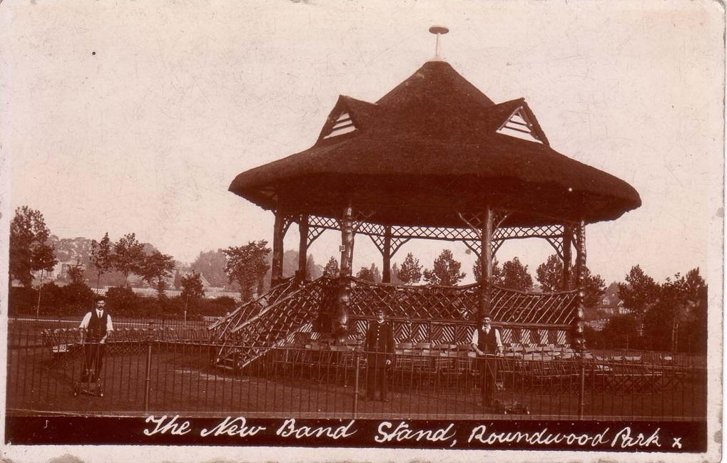 Black and white photograph of a wooden bandstand, with three men standing in front. Two men are wearing shirts and waistcoats and holding mechanical lawn mowers in front of them. One man is in park attendant uniform, with a black jacket and hat.