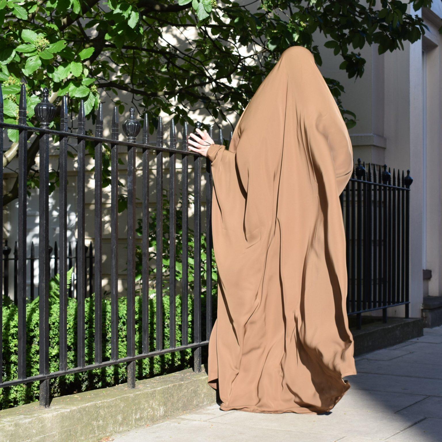 Colour photograph of woman wearing a jilbab in a natural brown tone. The photograph is taken from behind her and she is walking away from the camera along a London street, with wrought iron railings next to her.