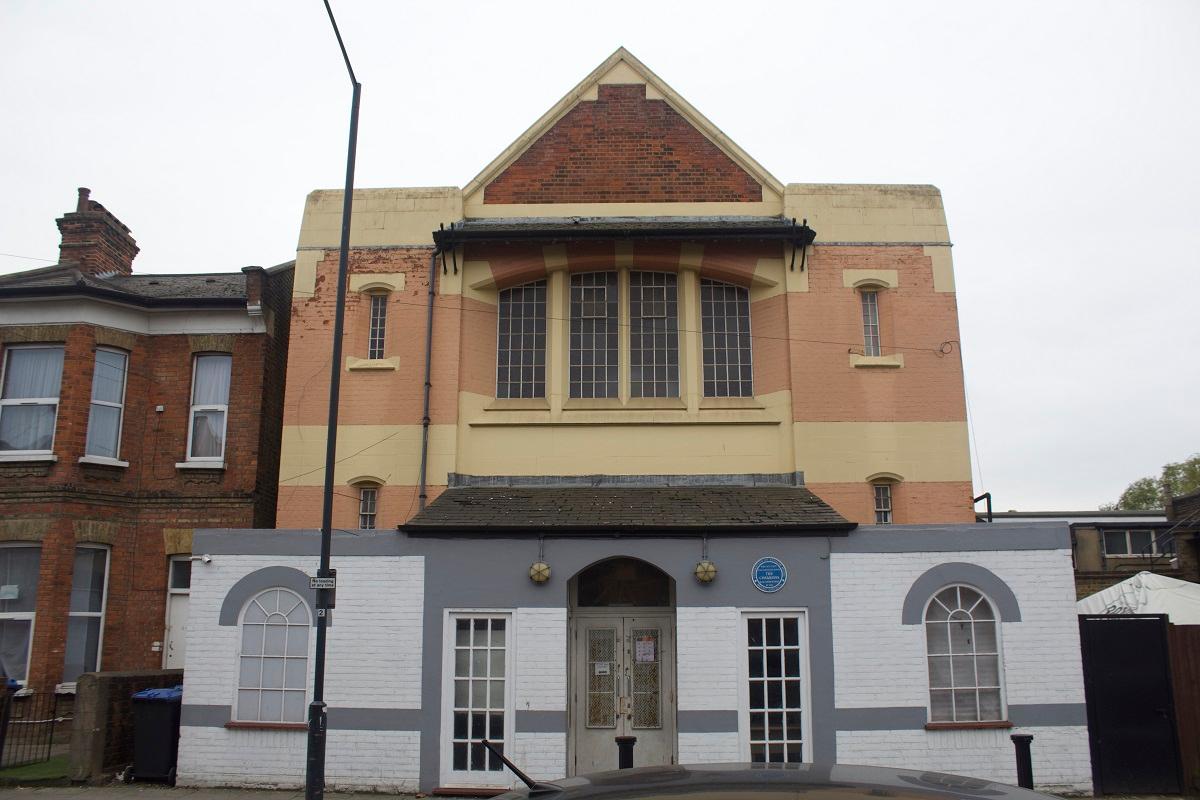 Colour photograph showing the front of a church hall with a blue plaque fixed to one side. The hall is red brick with a symmetrical design and large windows in the centre.