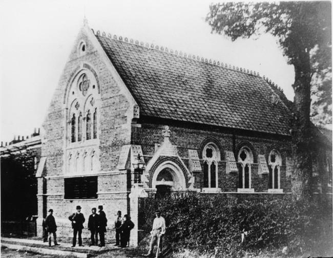 Black and white photograph of a small church with a group of men and boys standing outside. The church has made from bricks with ornate windows, but no spire.