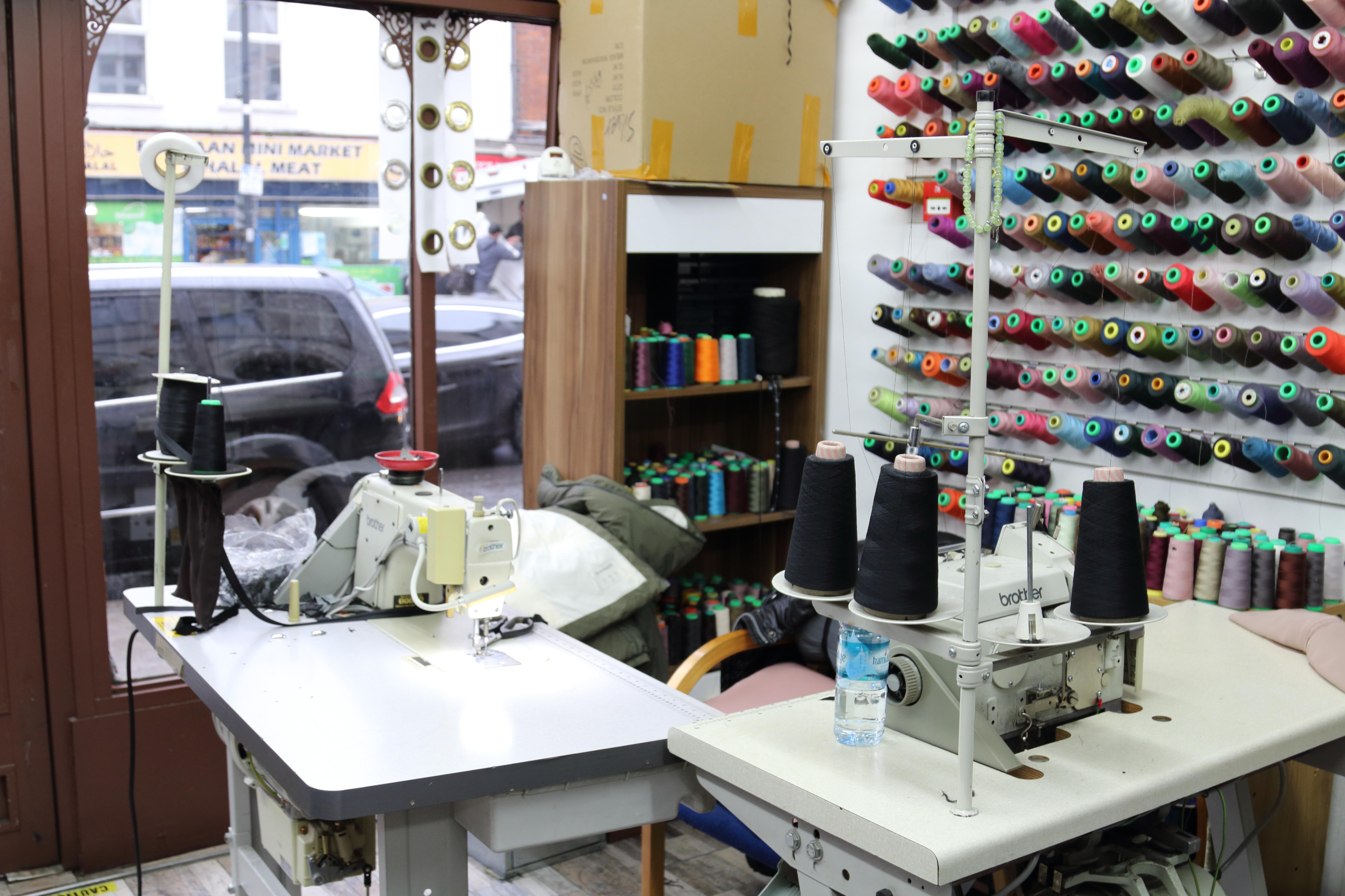 Photograph showing a corner of Ahmed Tailor’s shop where a tailor sits, with a work station including two different sewing machines. Behind the work station on the wall are spools of thread in different colours, in easy reach for use during day-to-day work.