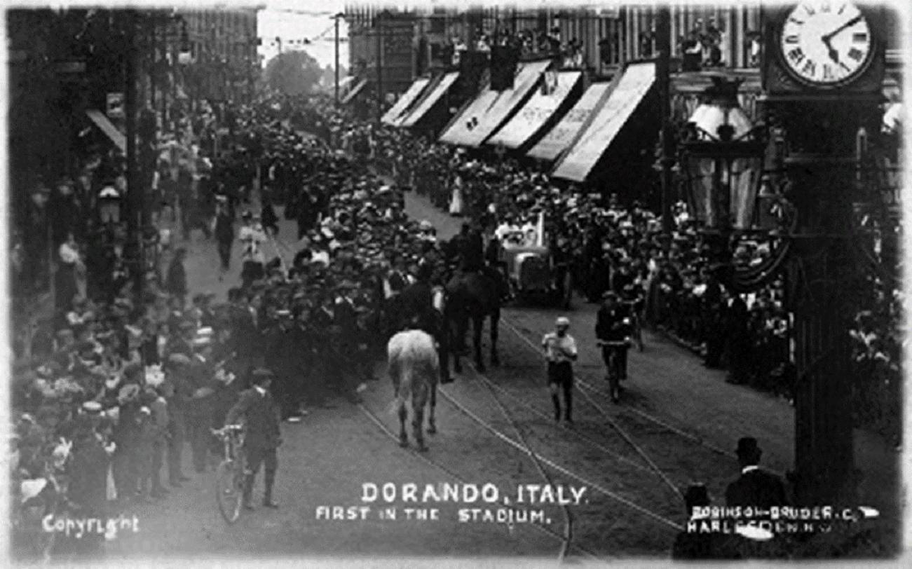 Postcard with a black and white photograph showing a street packed with crowds lining a route and a runner, a motor car and a police horse in the centre. An historic clock is in the foreground on the right-hand side. The card is annotated, ‘Dorando, Italy, first in the stadium’.