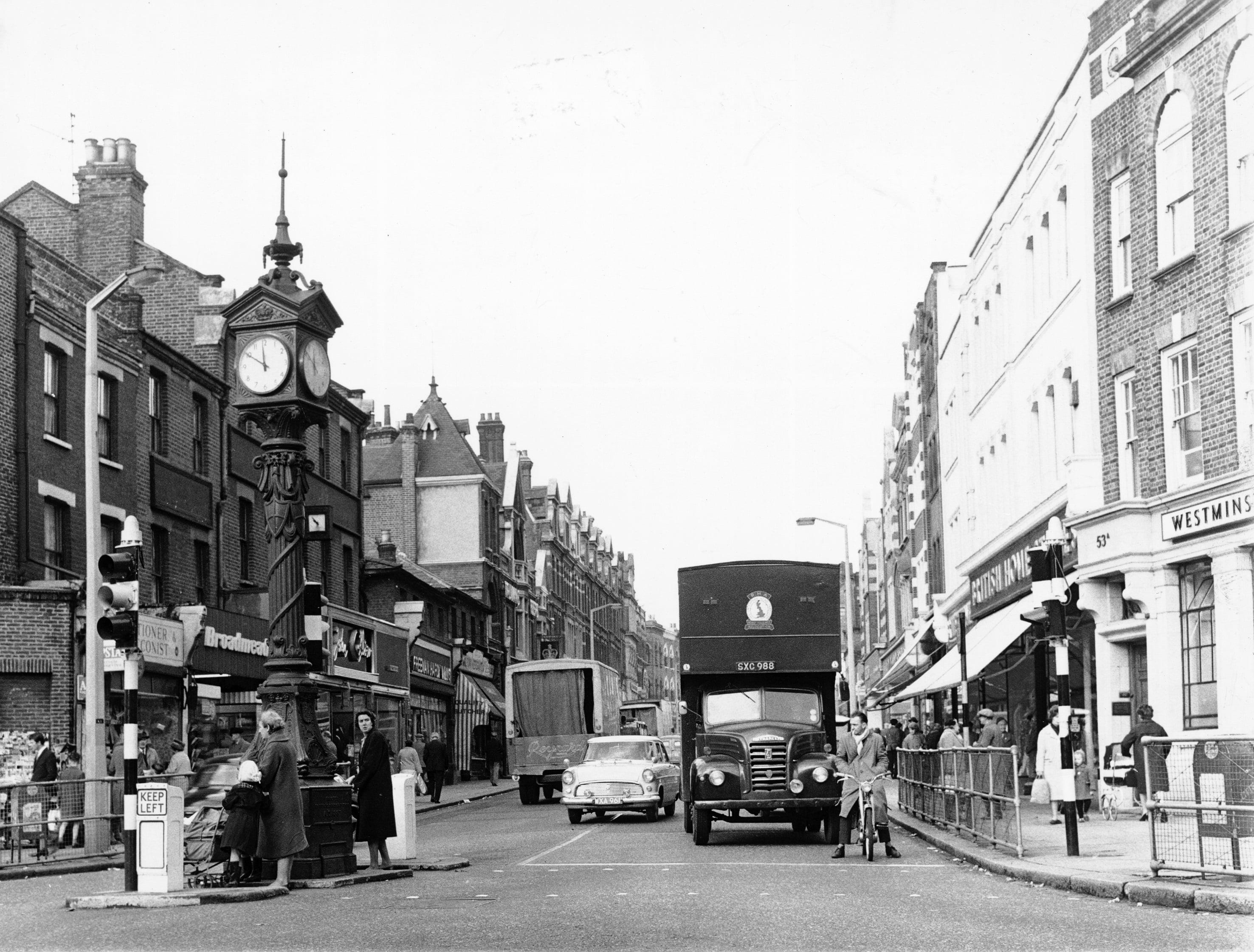 Black and white photograph showing a London street junction with busy traffic. An historic clock visible on the left hand side.