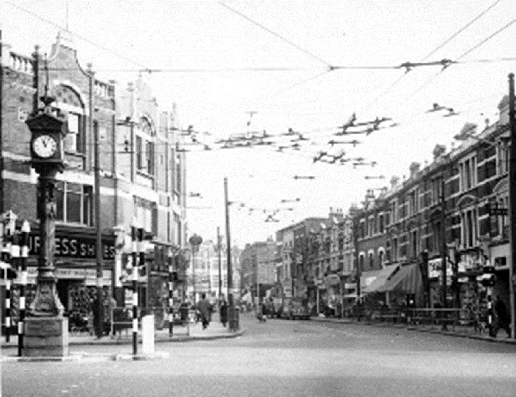 Black and white photograph showing a London street junction with tram wires overhead. An historic clock visible on the left hand side.