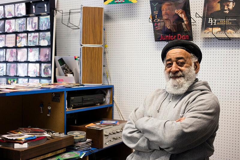 Colour photograph showing record shop owner Cleveland ‘Popsy’ Deer, of Starlight Records, behind his shop counter. There are records on the wall behind him. He is wearing a grey hooded sweatshirt, a black hat, and he has a grey beard. He is smiling, with his arms folded.