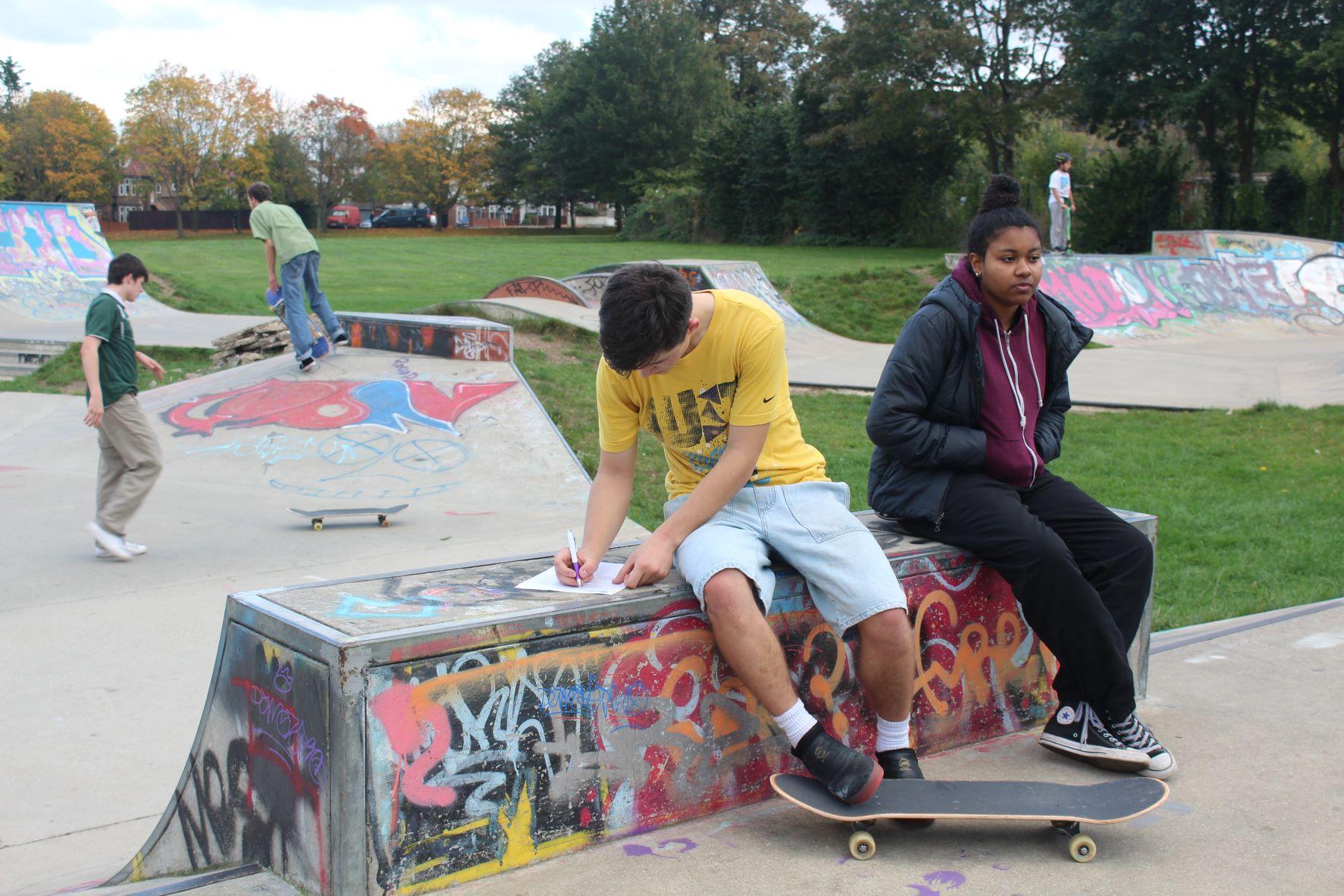 Colour photograph showing Roundwood Skatepark. In the foreground a young woman sits on a skate ramp next to a young man with a skateboard, writing on a piece of paper. In the background are two other young men, skating.