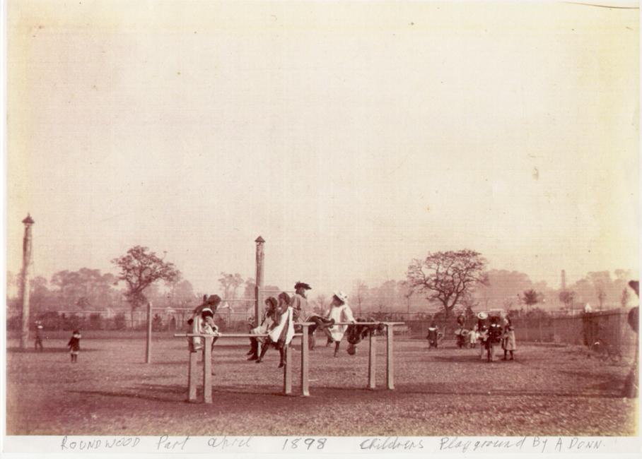 Sepia tinted black and white photograph showing young children climbing on a climbing frame in an open park space. Annotation reads: ‘Roundwood Park April 1898. Children’s Playground by A. Donn