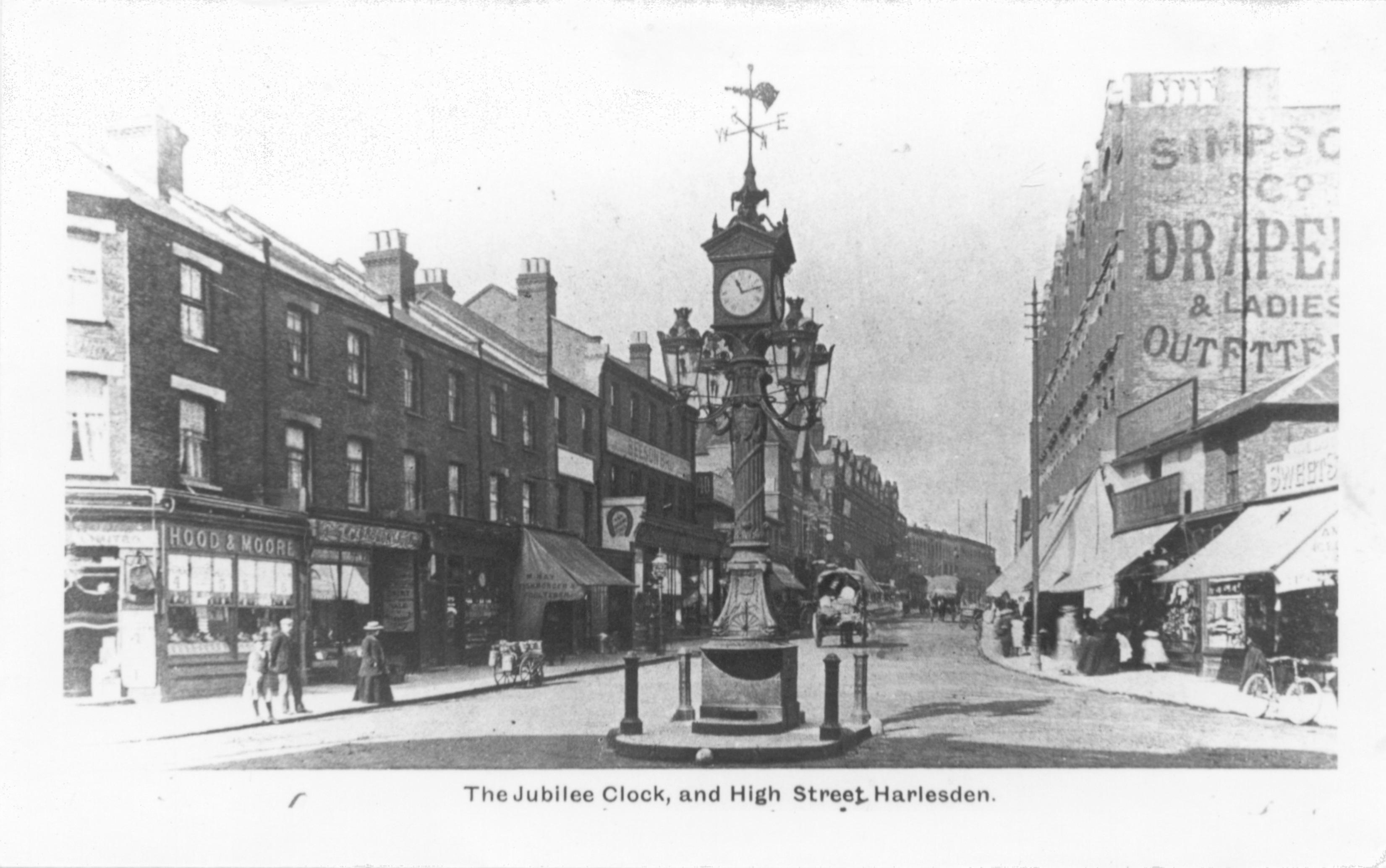 Black and white photograph showing a London street junction. An historic clock is in the centre of the junction. The shops have awnings and people are visible wearing dress from around the 1900s.