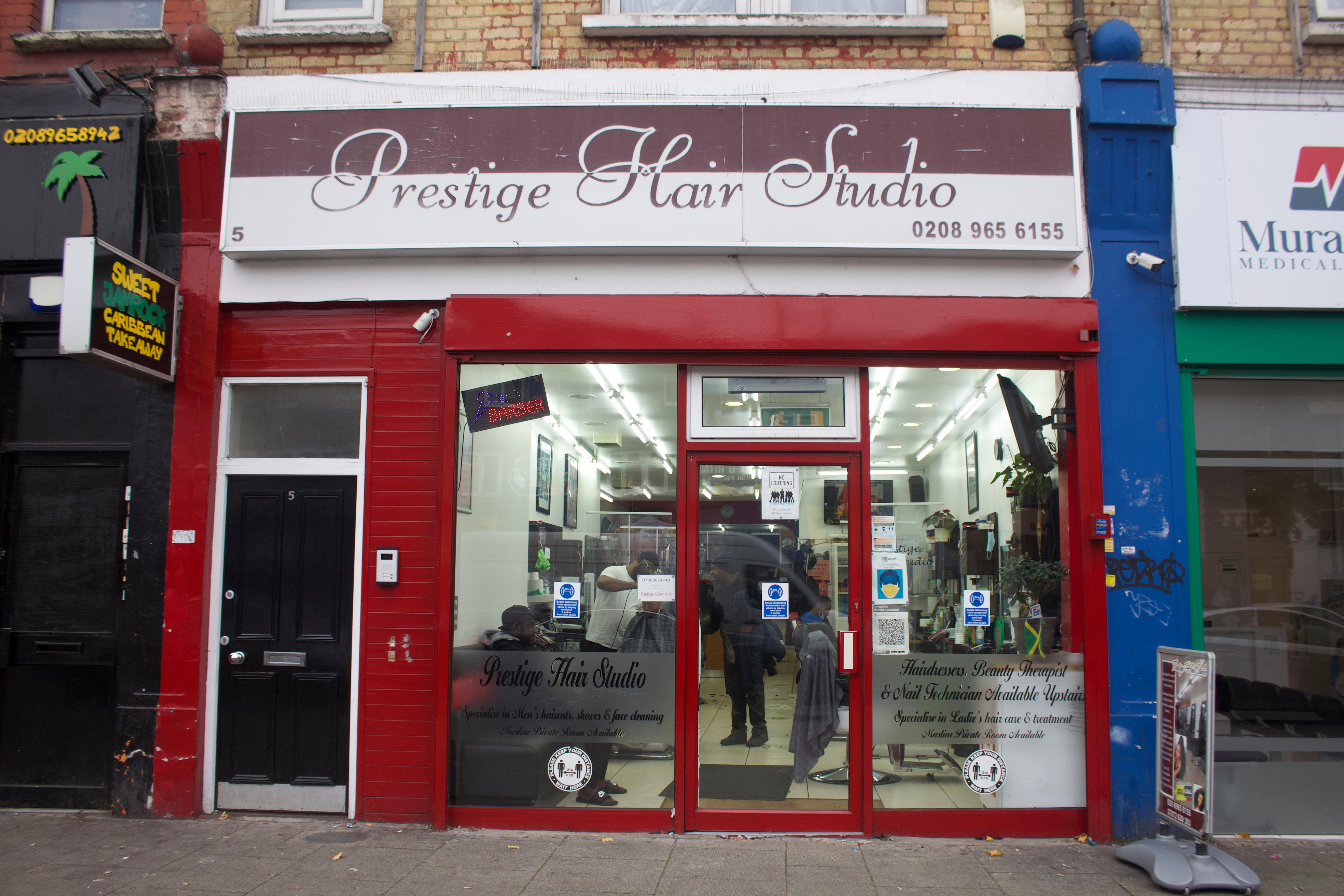 Shop front of a hairdressers, titled, ‘Prestige Hair Studio’. The lights in the shop are on and hairdressers are at work with customers.
