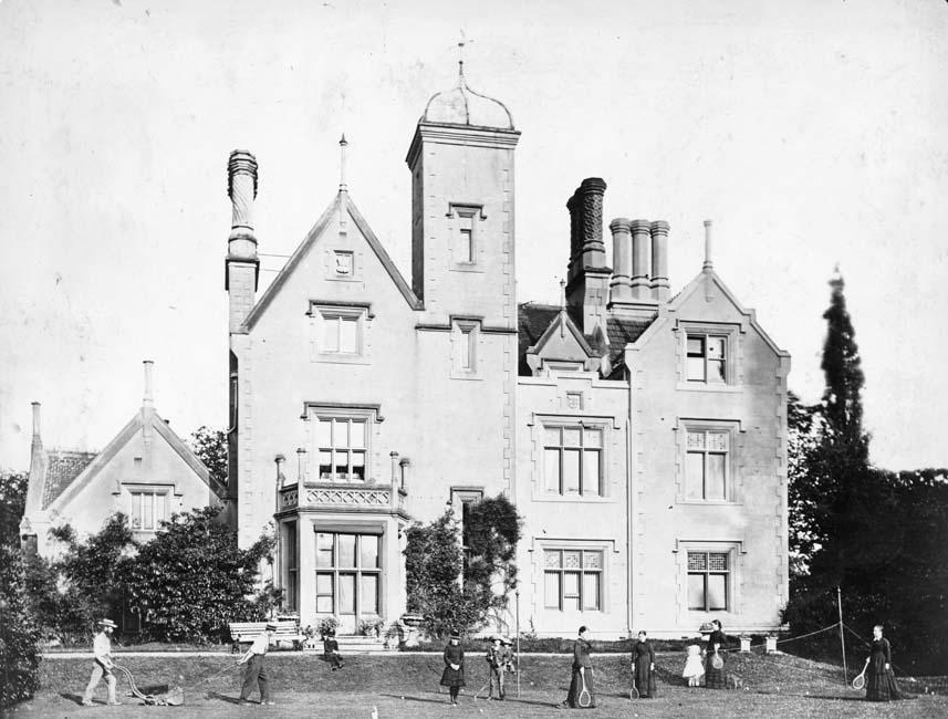 Black and white image of a large victorian house with tower and decorated chimneypots. In the foreground is a group of women and children playing tennis or badminton. On the left hand side are two men, possibly servants, mowing the lawn.