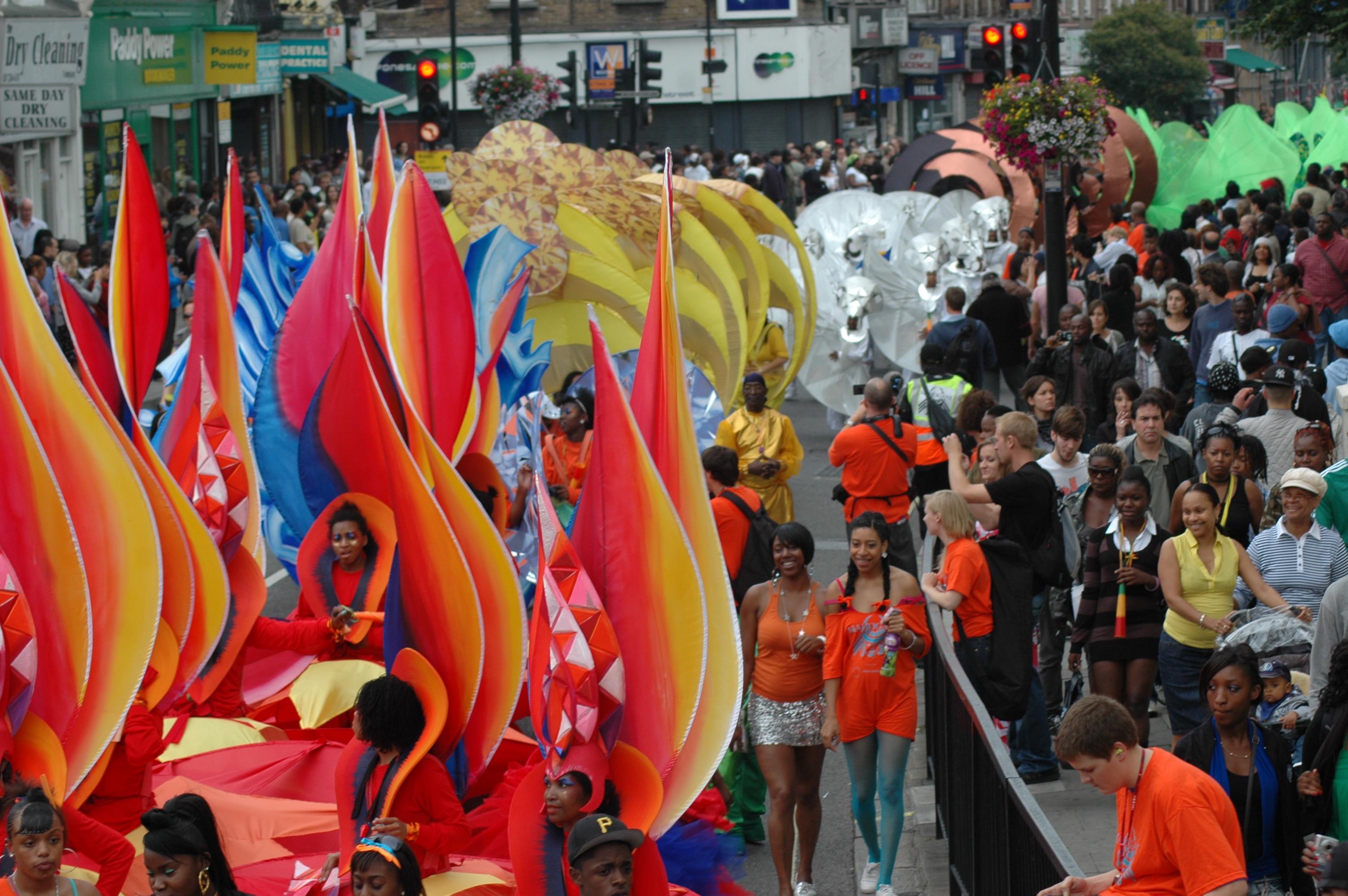 Colour photograph of a street carnival with elaborate red and orange costumes in the foreground. Spectators can be seen smiling and walking alongside the parade, behind a barrier, on the right-hand side.