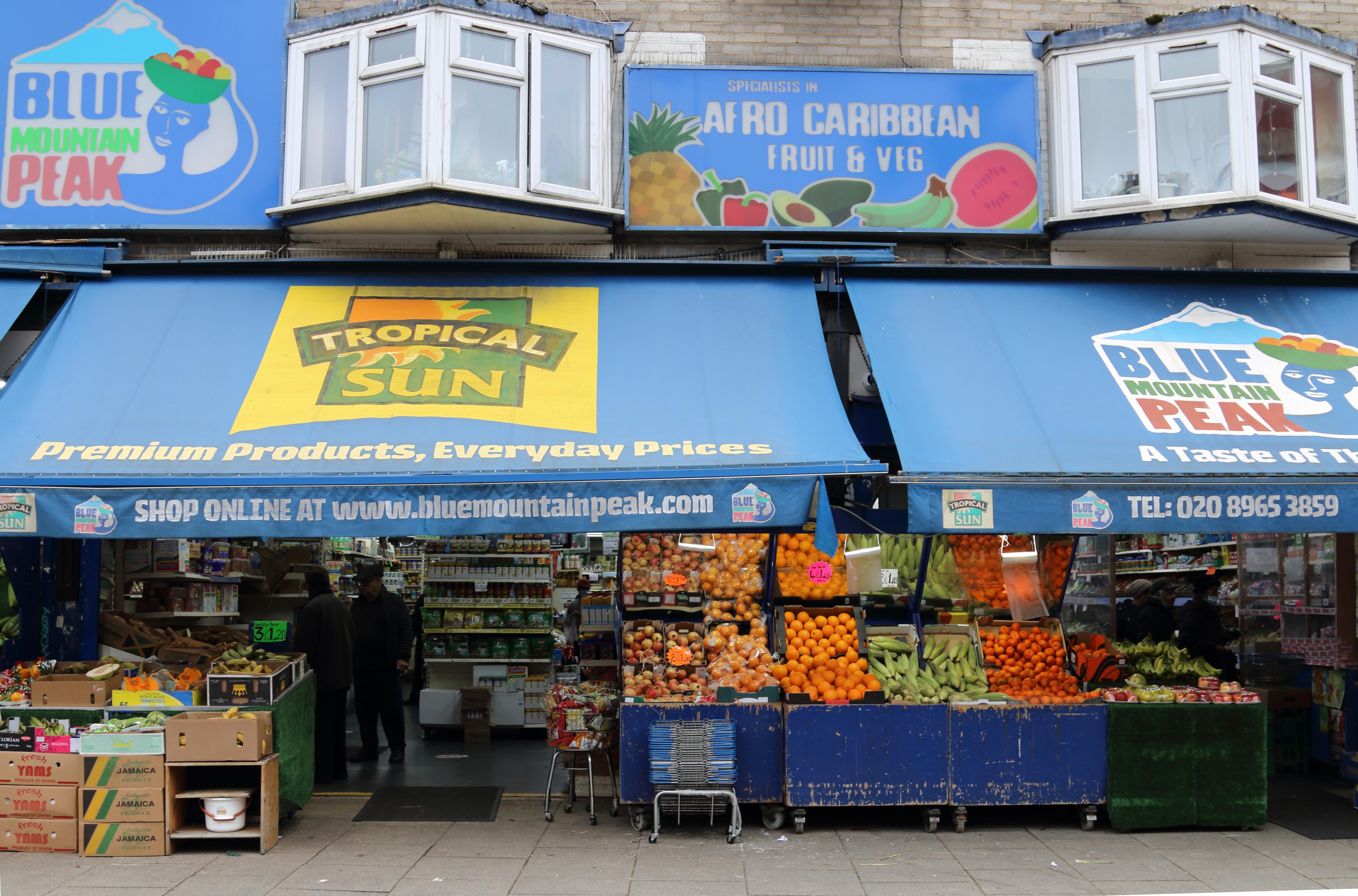 Shop front of a grocery store with fruit displayed at the entrance. The shop has blue awnings and the shop sign reads, ‘Blue Mountain Peak’ and ‘Afro-Carribean Fruit & Veg’