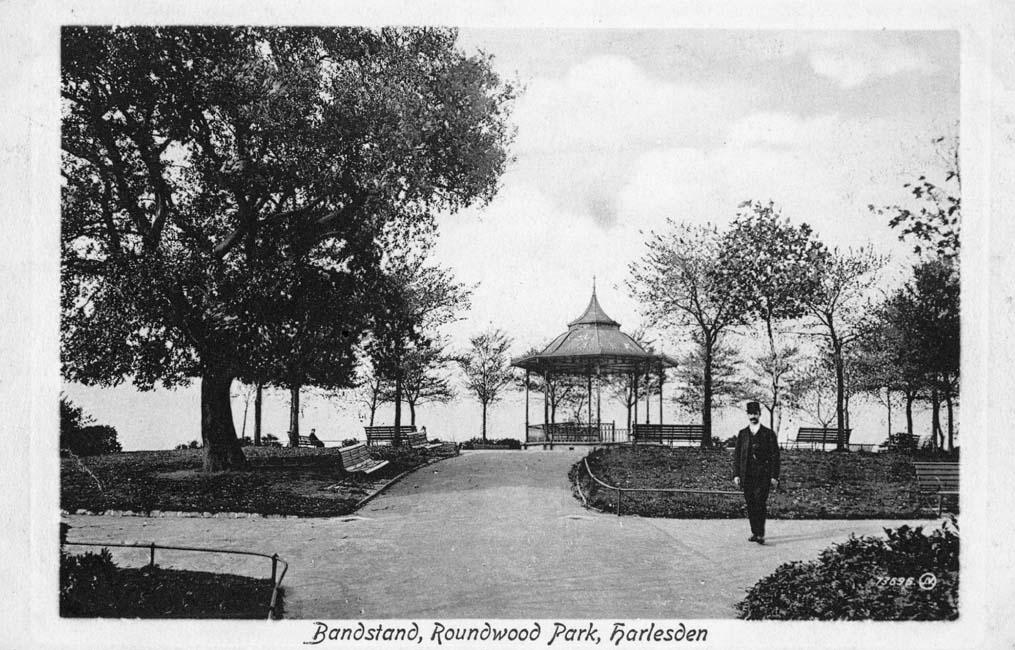 Black and white photograph showing a park area with a bandstand in the background. In the foreground stands a park attendant in uniform.